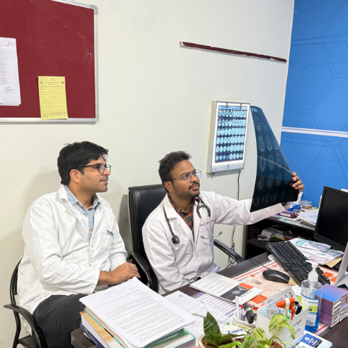Two male doctors in white coats examine a chest X-ray film held up by one doctor in a cluttered clinic office.