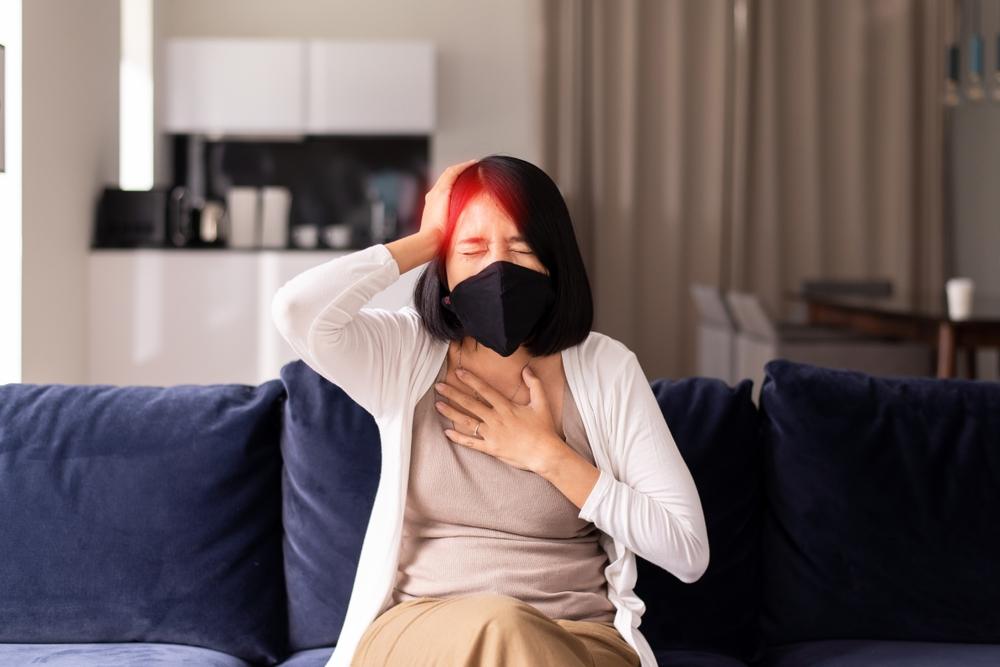 Woman with a black face mask sits on a blue sofa, one hand on her chest and the other on her head, looking distressed in a living room.