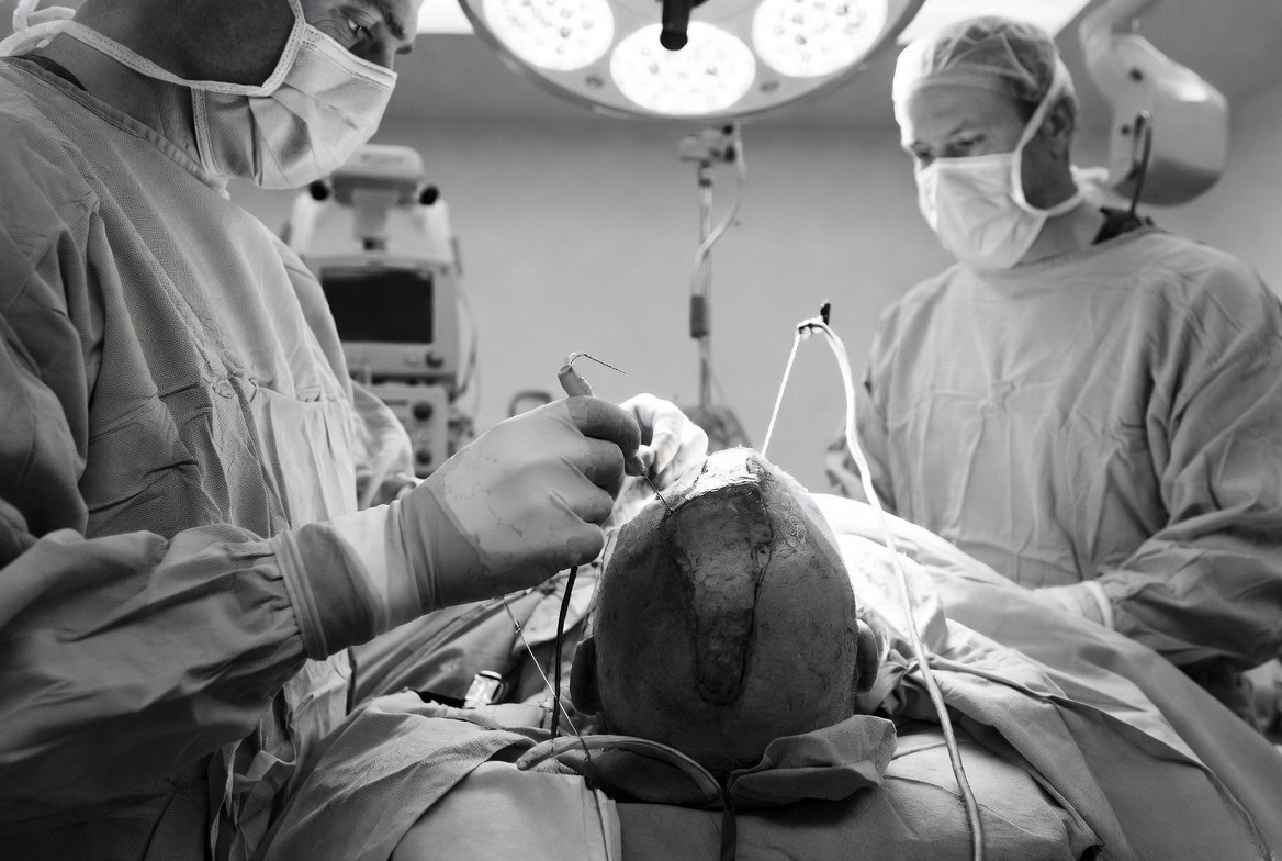 Two surgeons in masks perform brain surgery, focusing on the patient’s head with instruments under bright overhead lights in an operating room.