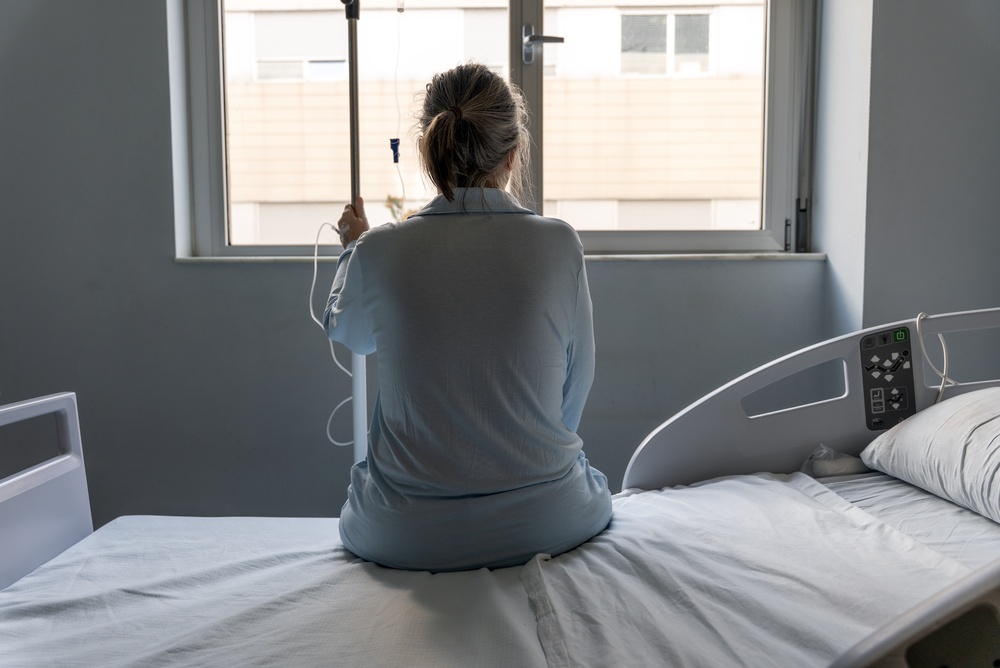 Person in a blue hospital gown sits on the edge of a bed, facing a window with an IV pole in hand (back view).