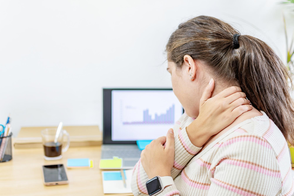 Woman at a desk holds her neck in pain while looking at a laptop displaying charts and graphs.